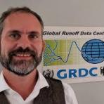 A man with a beard and wearing a white shirt and vest stands in front of a sign that reads "Global Runoff Data Centre" and "GRDC" with a globe and river graphic.