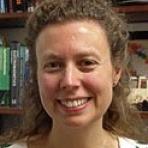 Smiling person with curly hair stands in front of a bookshelf filled with books and decorative items.